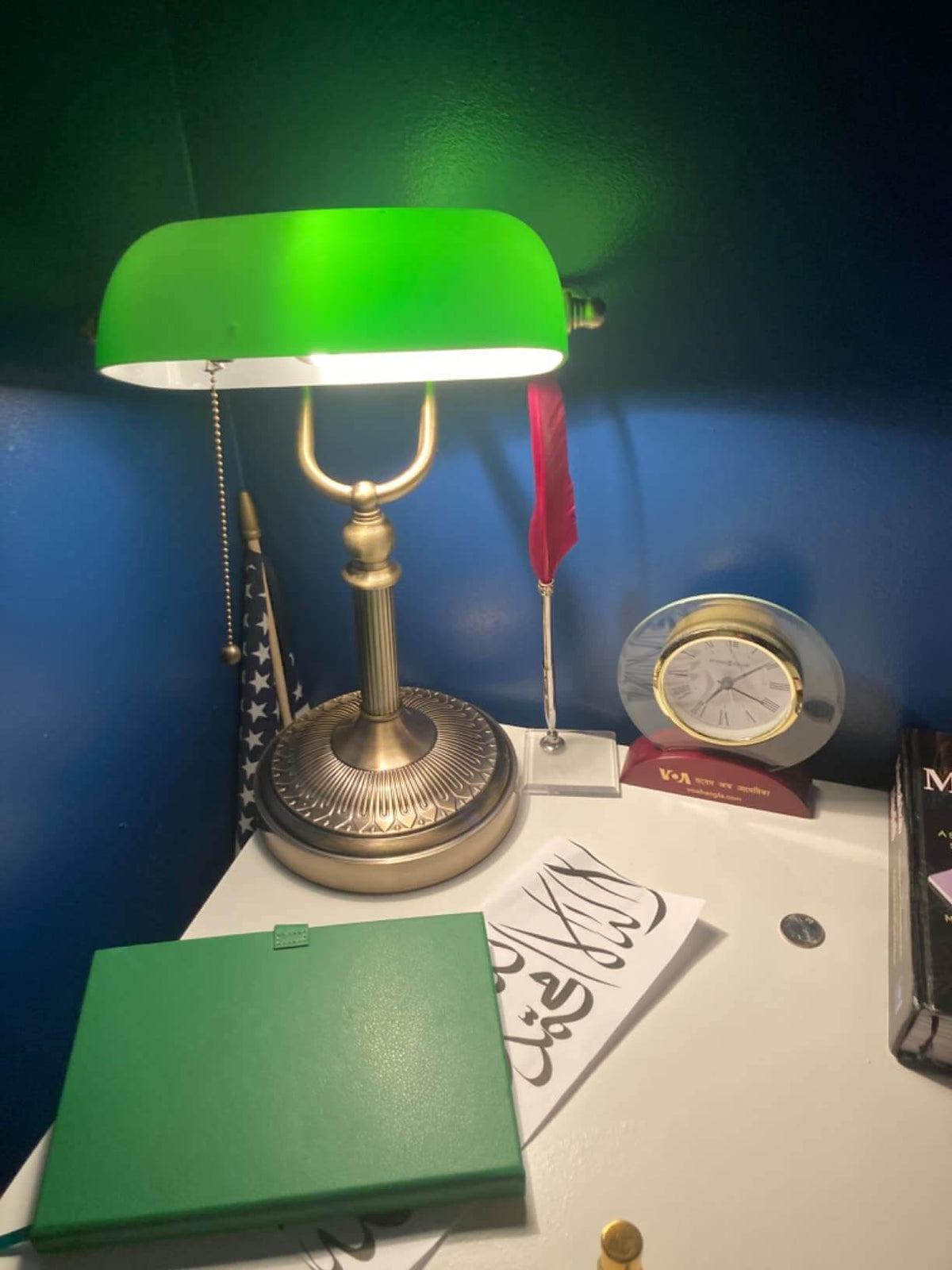 Newrays Green Glass Banker Lamp on desk with a green notebook and clock, complementing a blue wall background.