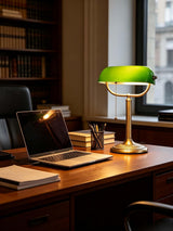A classic green glass bankers desk lamp illuminating a workspace with a laptop and books.
