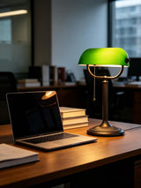 Antique green glass bankers desk lamp beside a laptop on a wooden desk in a well-lit office setting.